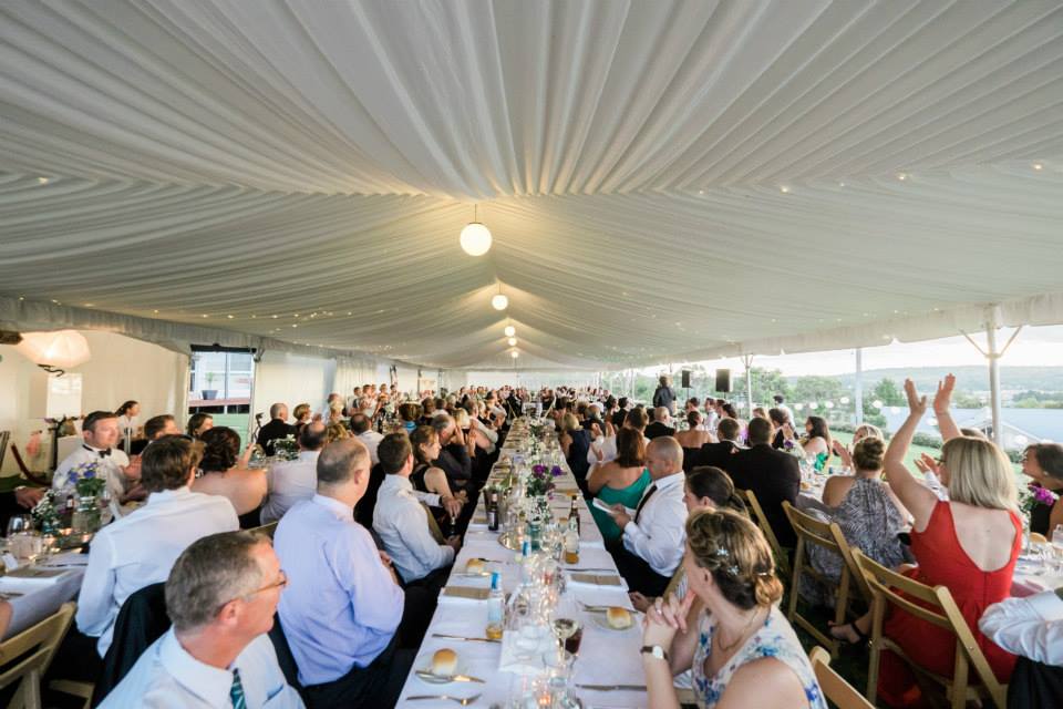 A large group of people are sitting at long tables under a tent