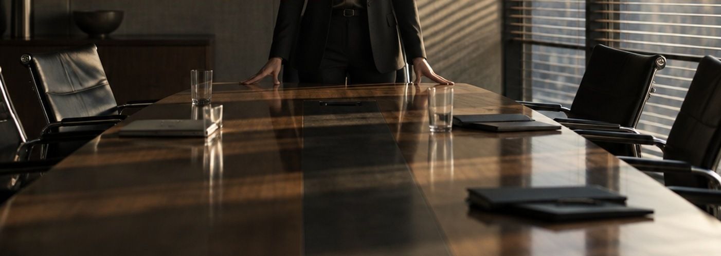 A woman standing at the head of an empty boardroom table, hands flat on the wood