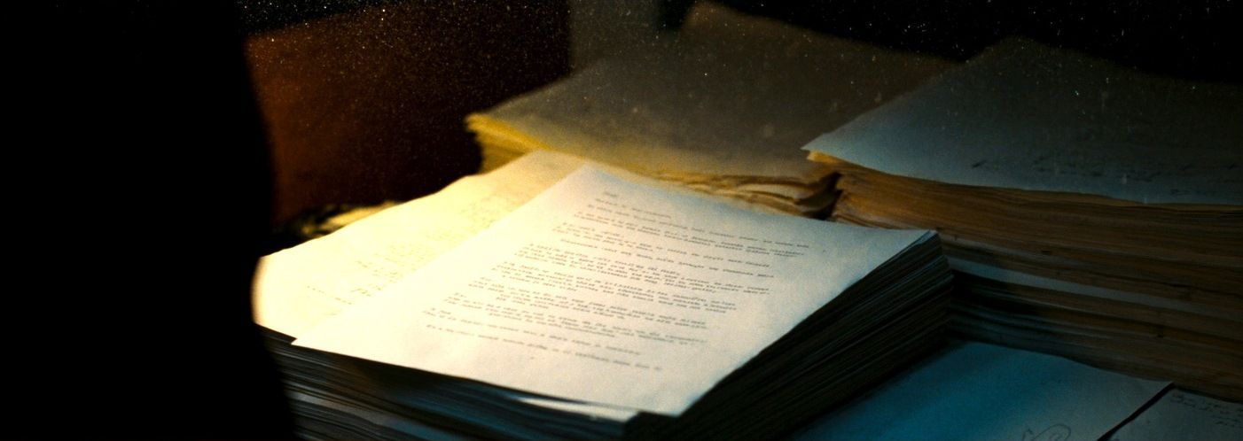 A single screenplay page on top of a tall stack of older yellowed drafts under a desk lamp
