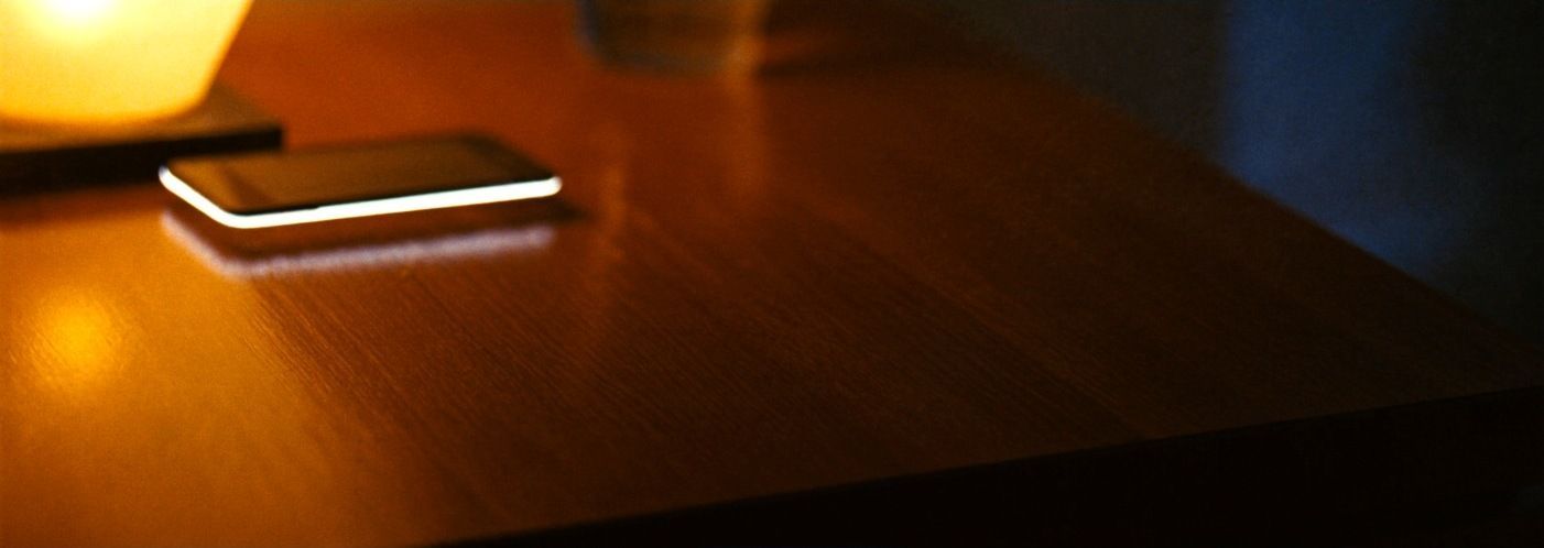 A phone lying face down on a bedside table at night, screen glow leaking around its edges
