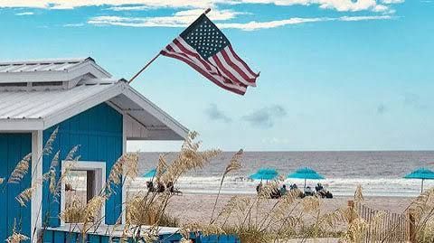 Beachside blue cottage with American flag, dune grass, and turquoise umbrellas by the ocean