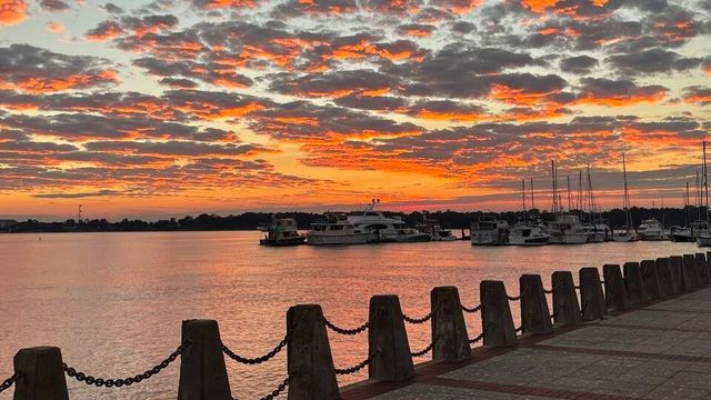 Sunset over a harbor with boats, orange clouds, and a waterfront walkway lined with posts and chains