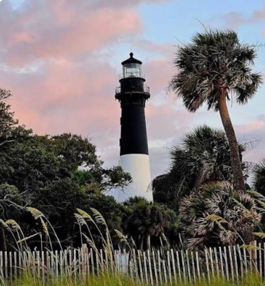 Black-and-white lighthouse above tropical trees and a fence at sunset with pink clouds