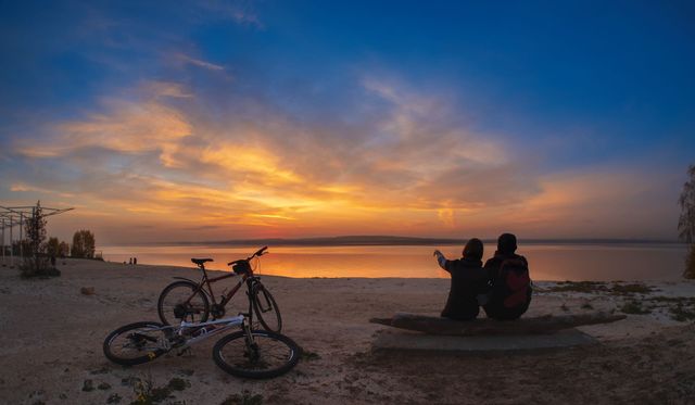 Bicycle by a beach at sunset, with two people sitting and looking at the horizon.
