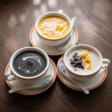 Three small cups of creamy desserts on saucers, topped with syrup and fruit on a wooden table.