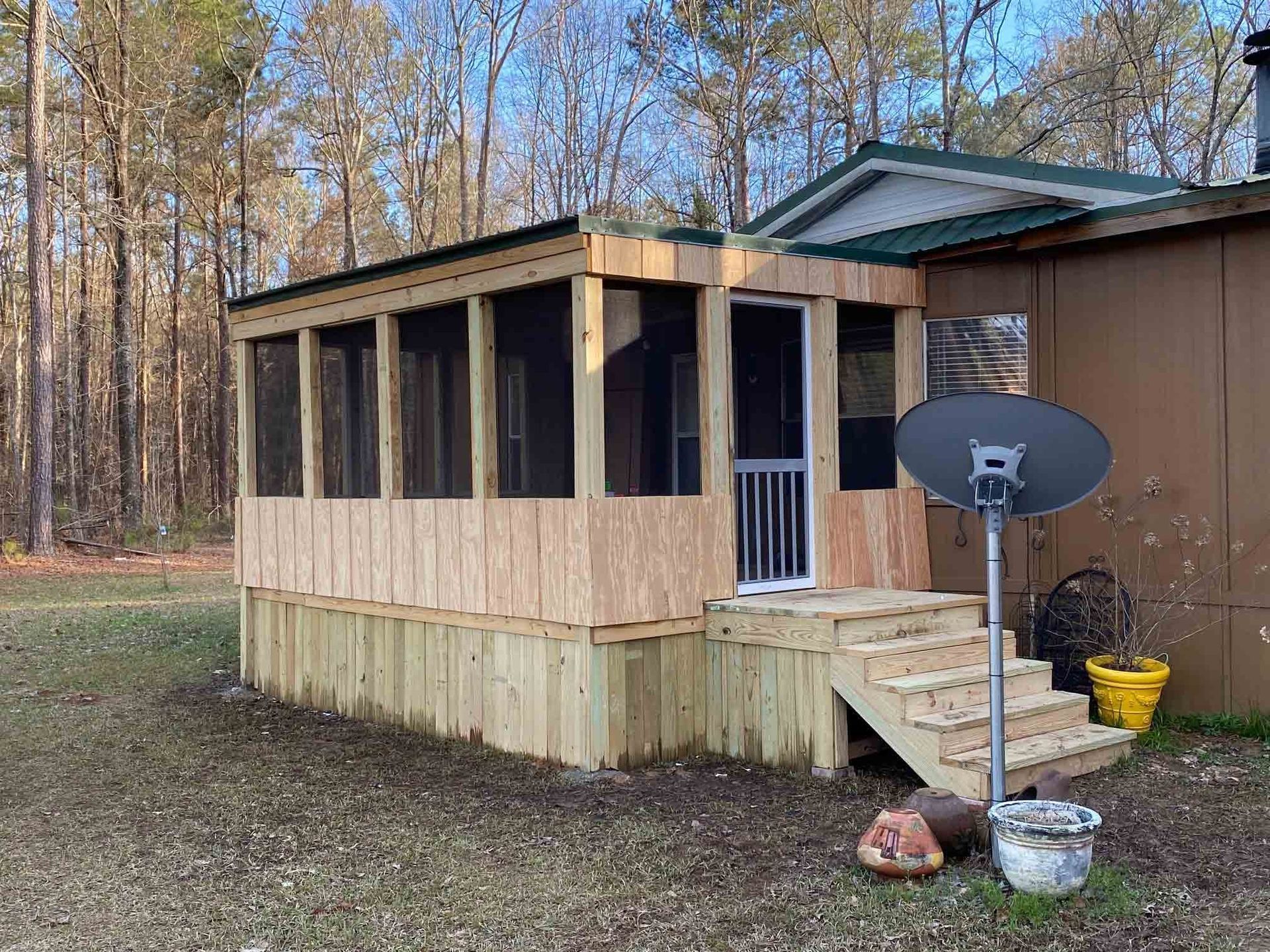 A newly constructed wood-framed screened porch with plywood siding and steps, attached to a brown house in a wooded area.