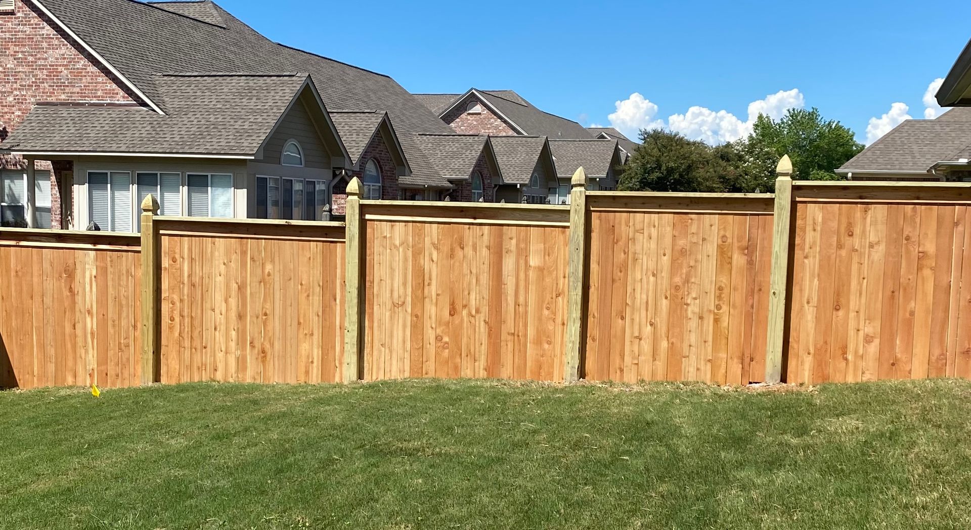 A newly installed light-colored wood privacy fence stands on a lush green lawn, extending toward a brick house corner.