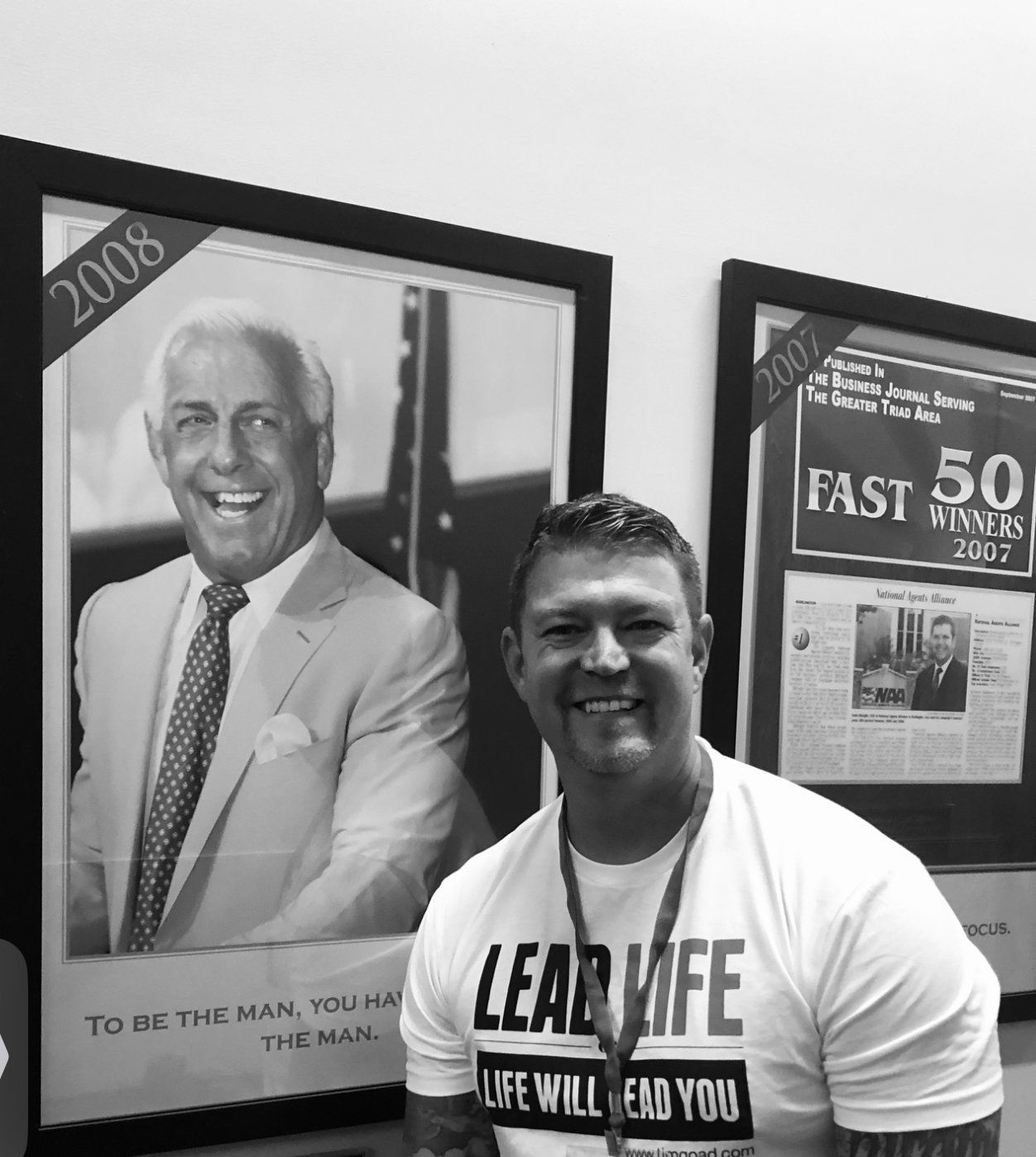 Man standing indoors beside framed magazine covers, smiling at the camera, wearing a white T-shirt and lanyard.