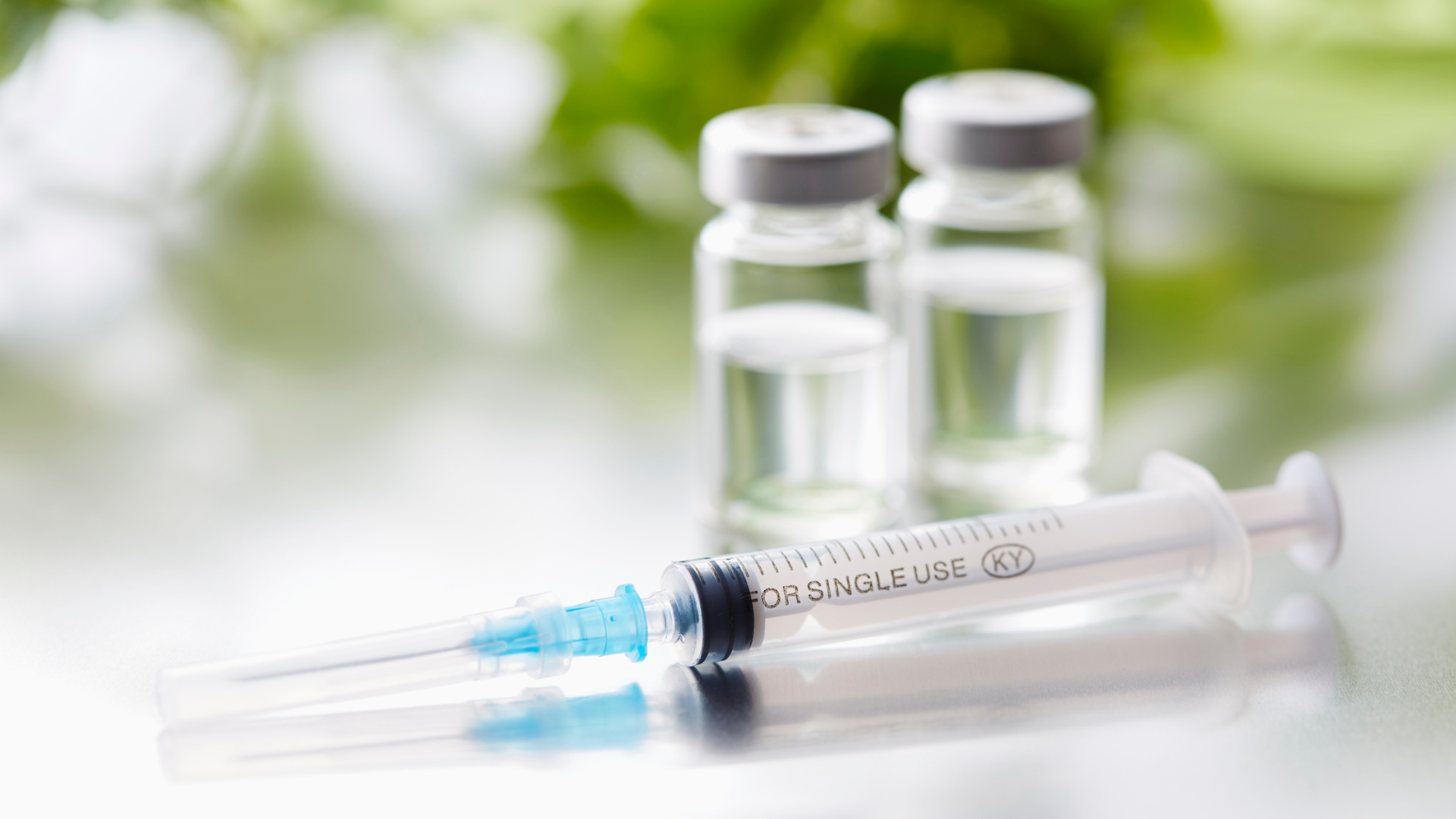 Two syringes and two medication vials on a white surface, with a soft green blurred background.