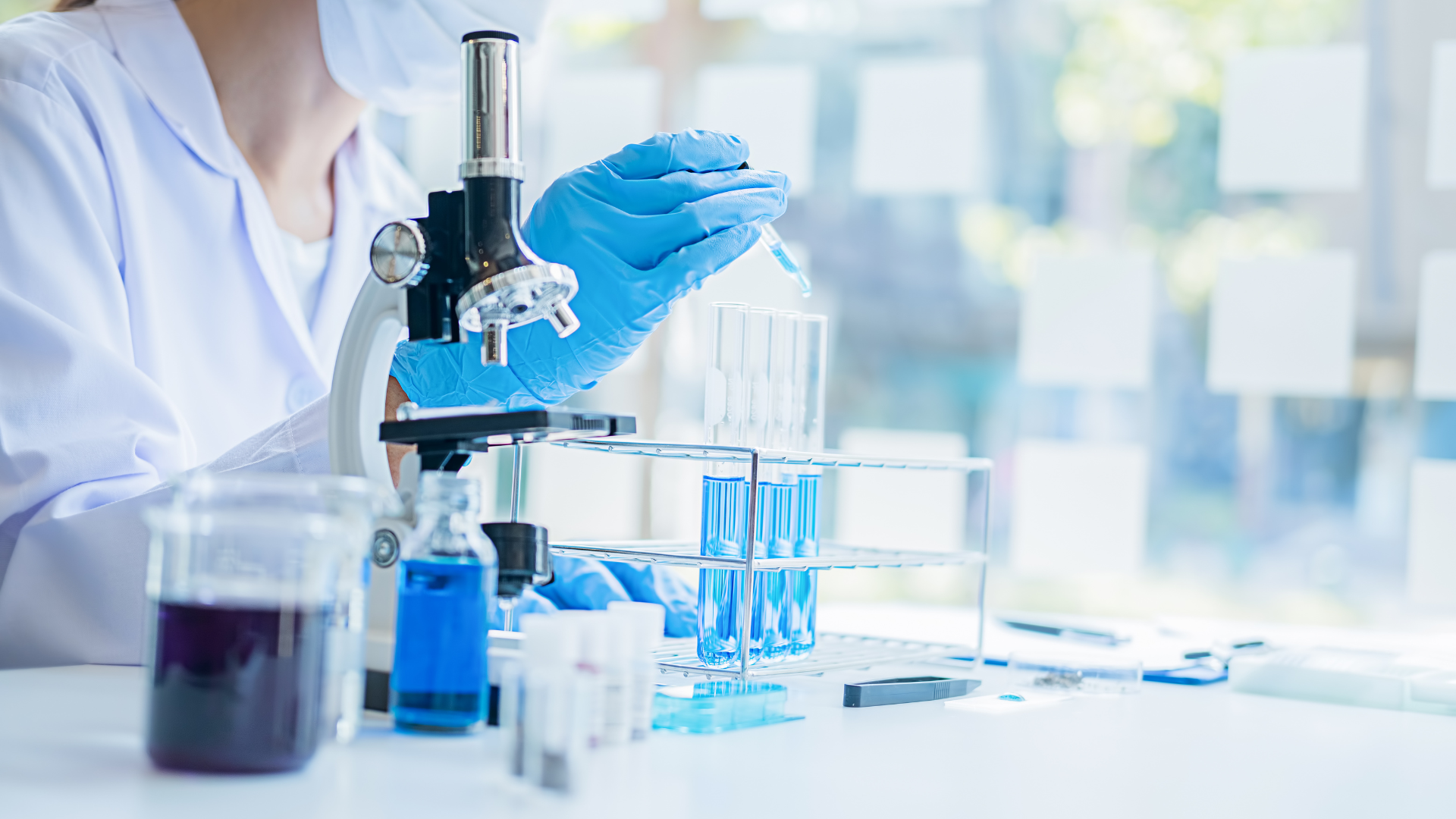 Gloved scientist using a microscope beside test tubes in a bright laboratory
