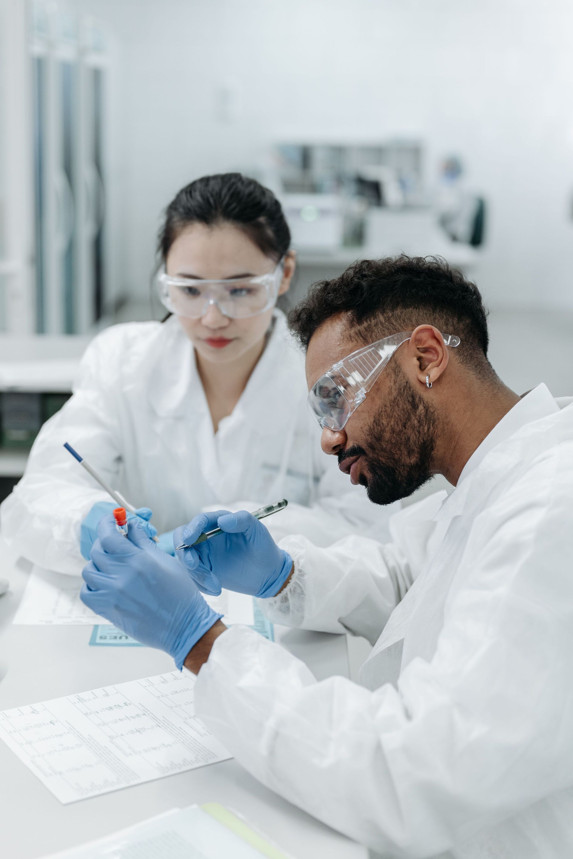 Two scientists in white lab coats and safety goggles examine a sample in a lab.