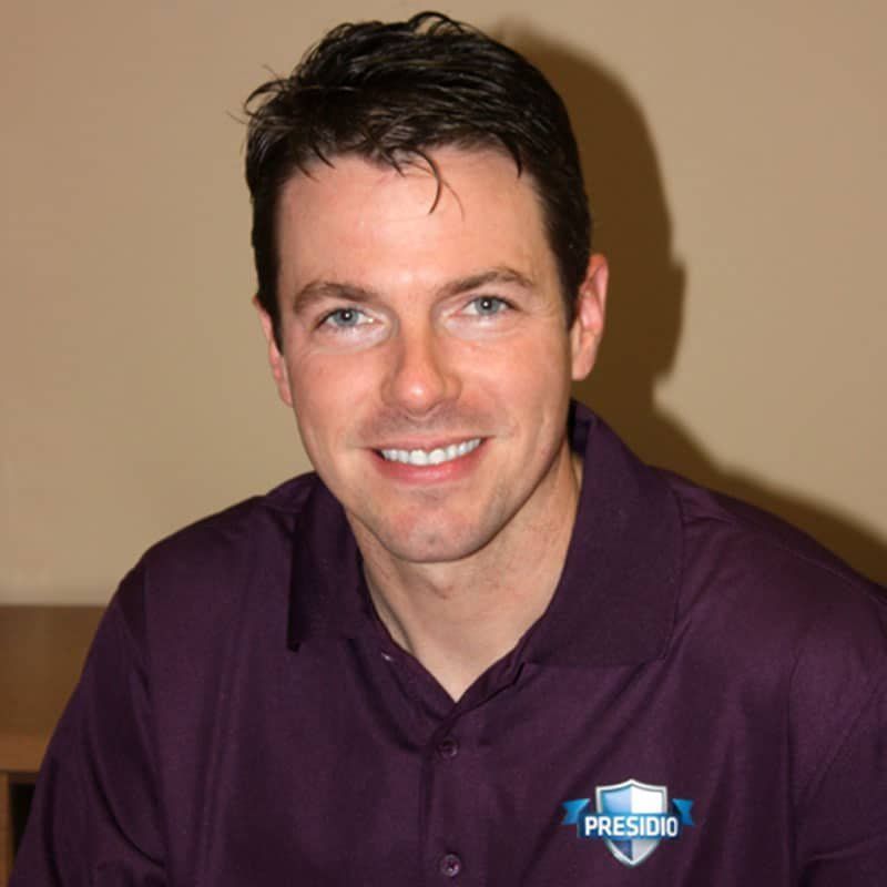 Man in a purple shirt smiling against a tan background, wearing a Presidio logo badge