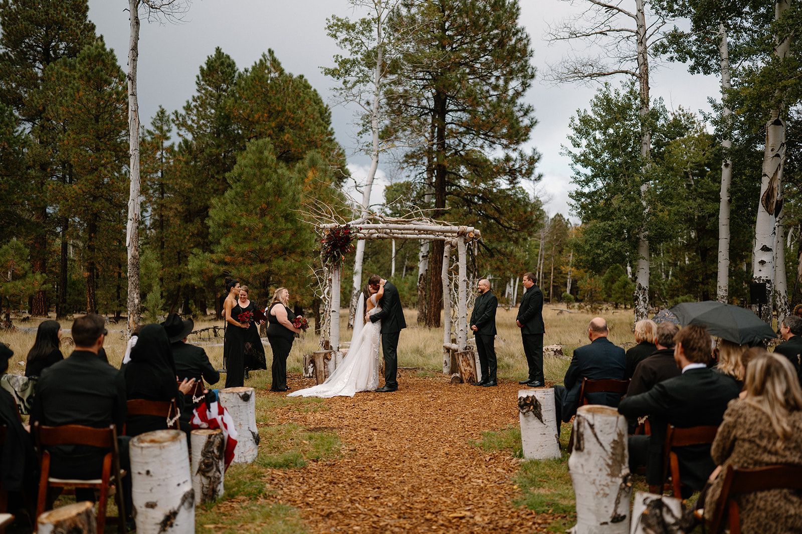 A wedding ceremony taking place in a forest clearing with guests seated on chairs made of birch logs.