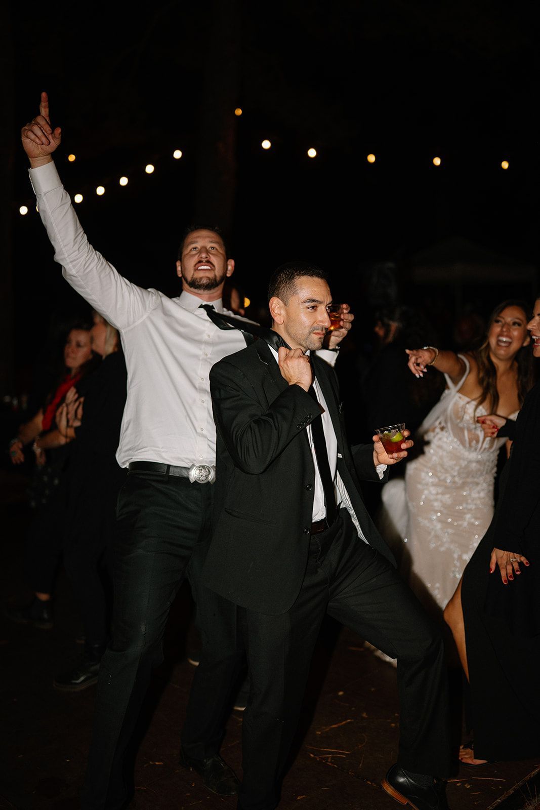 Two men in formal wear dancing at a lively wedding, one holding a drink while the bride watches.