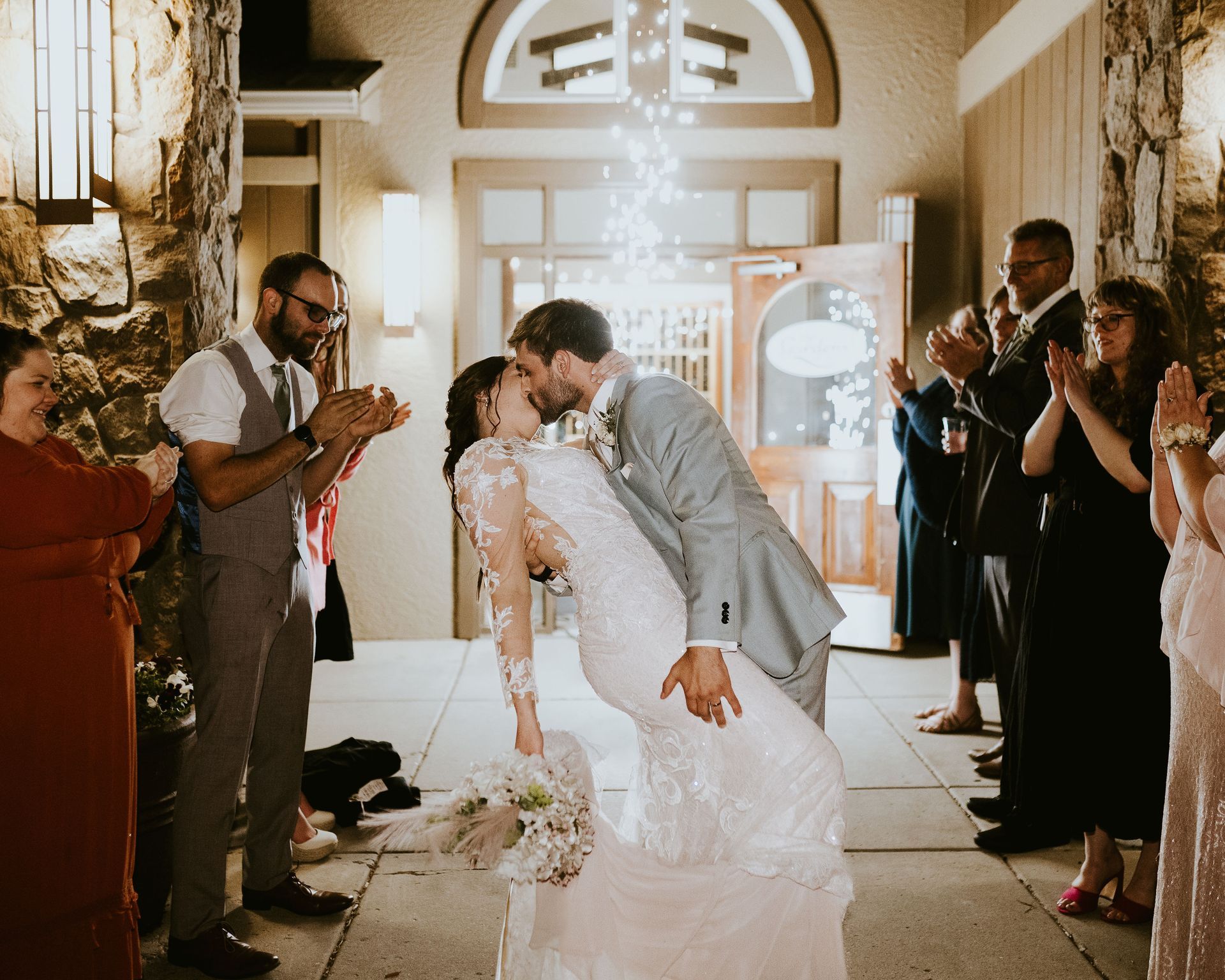 A newlywed couple kisses in a dip during their wedding exit as guests applaud in a stone-walled outdoor entryway.