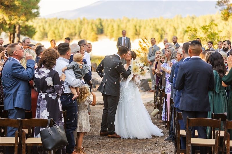 Bride and groom kiss, walking through guests throwing confetti after an outdoor wedding.