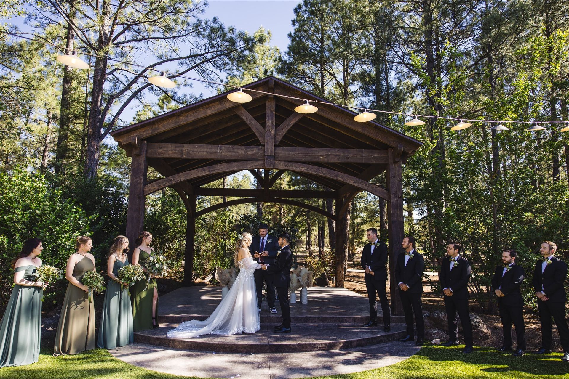 A bride and groom exchange vows under a wooden pavilion in a forest, surrounded by bridesmaids and groomsmen.