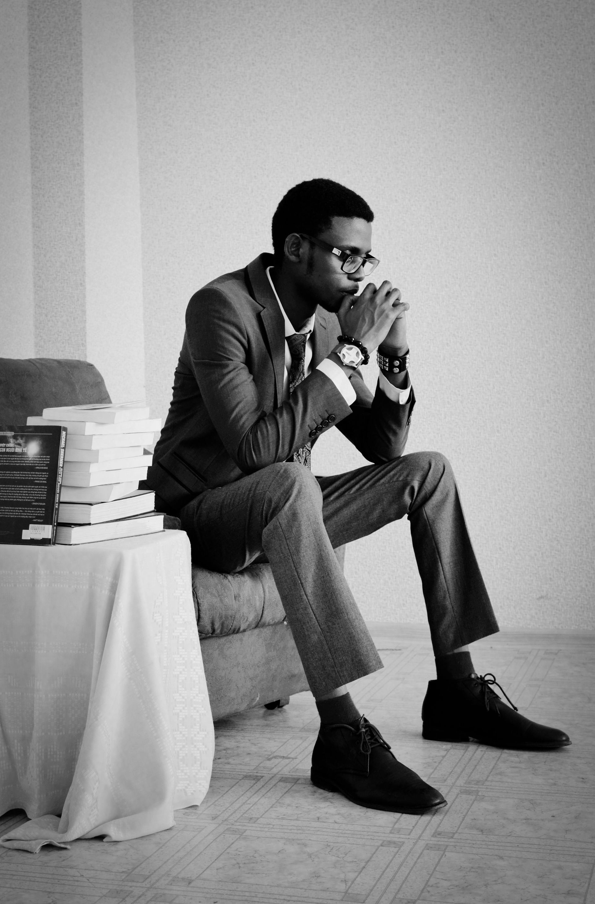A man in a suit is sitting on a couch in front of a stack of books.