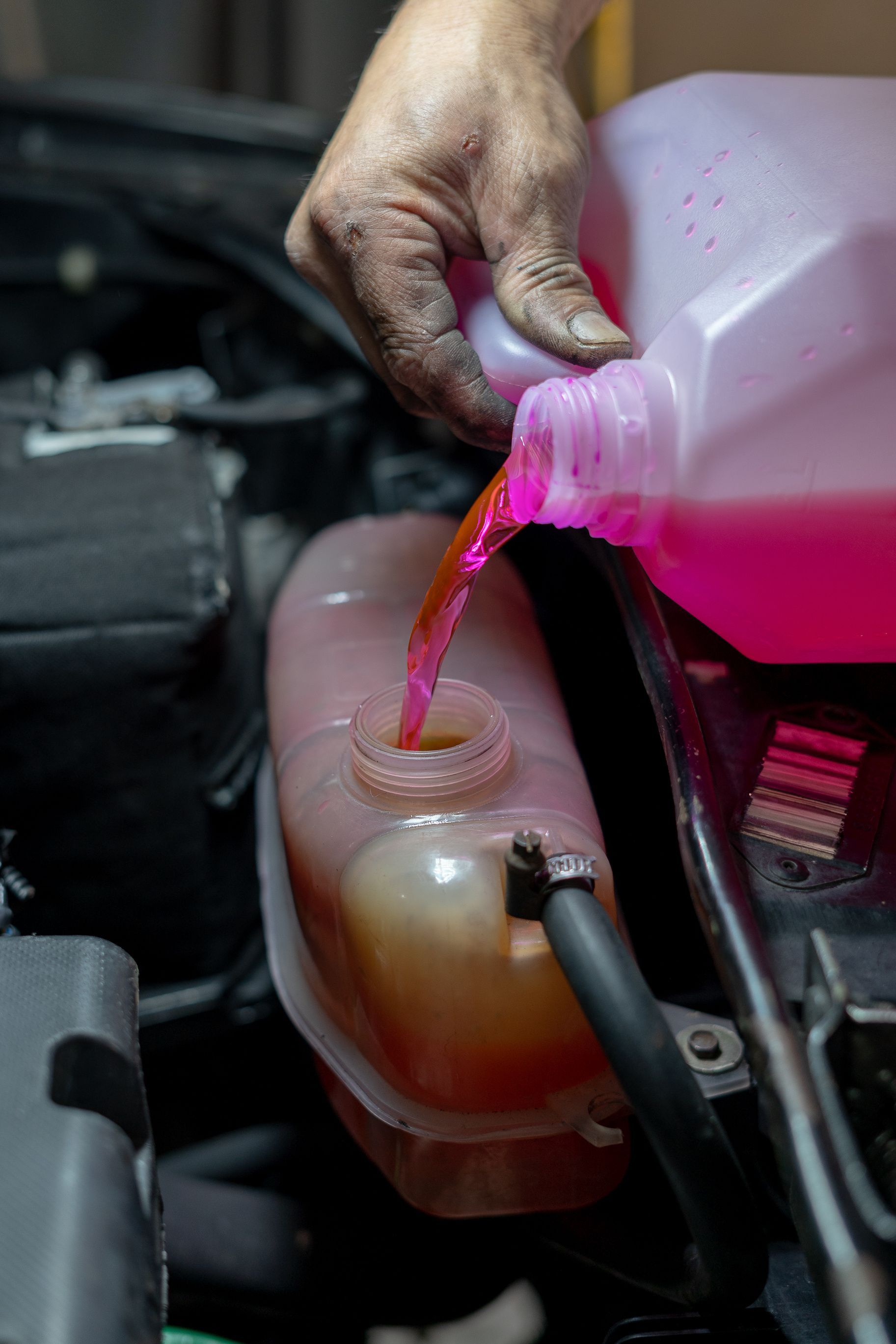 Hand pouring pink coolant into a car's reservoir. Dirty hand, black hose visible.