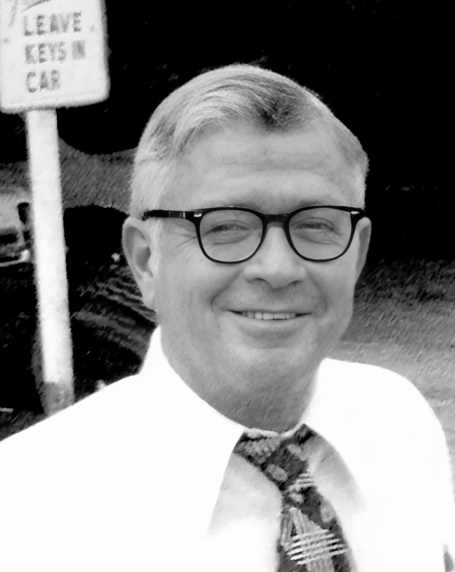 Man with glasses smiles, wearing a tie and white shirt. Sign