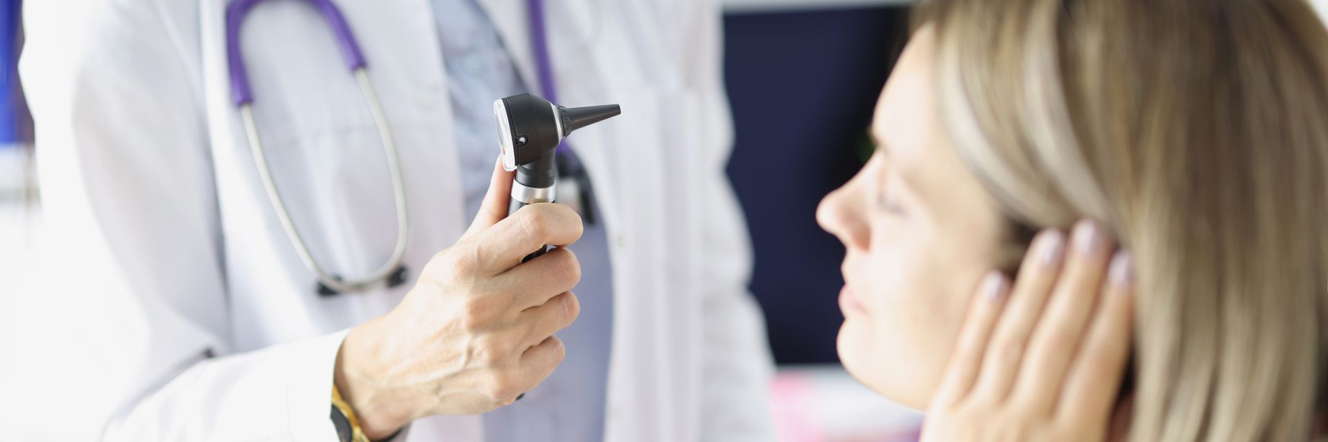 An audiologist holds an otoscope while examining a seated patient’s ear in a clinical exam room An audiologist holds an otoscope while examining a seated patient’s ear in a clinical exam room