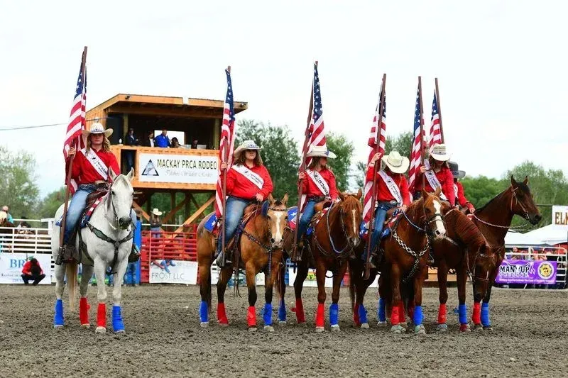 Six riders on horseback, holding American flags at a rodeo. Riders wear red sashes, blue and red leg wraps.