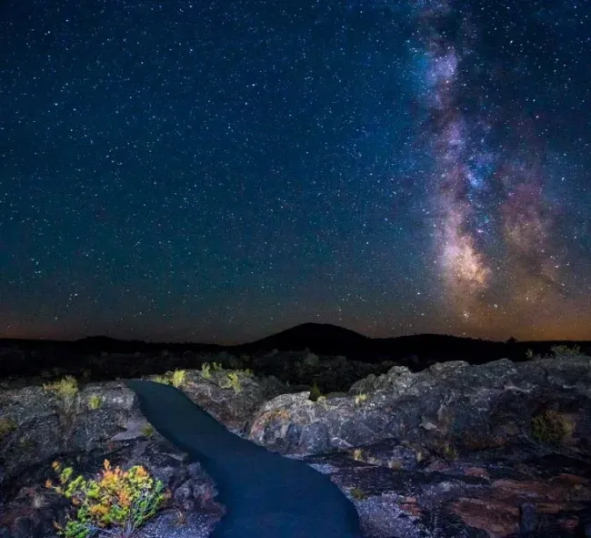 Dark starry sky over a dark, rocky landscape with a paved path leading toward the horizon. Milky Way visible.