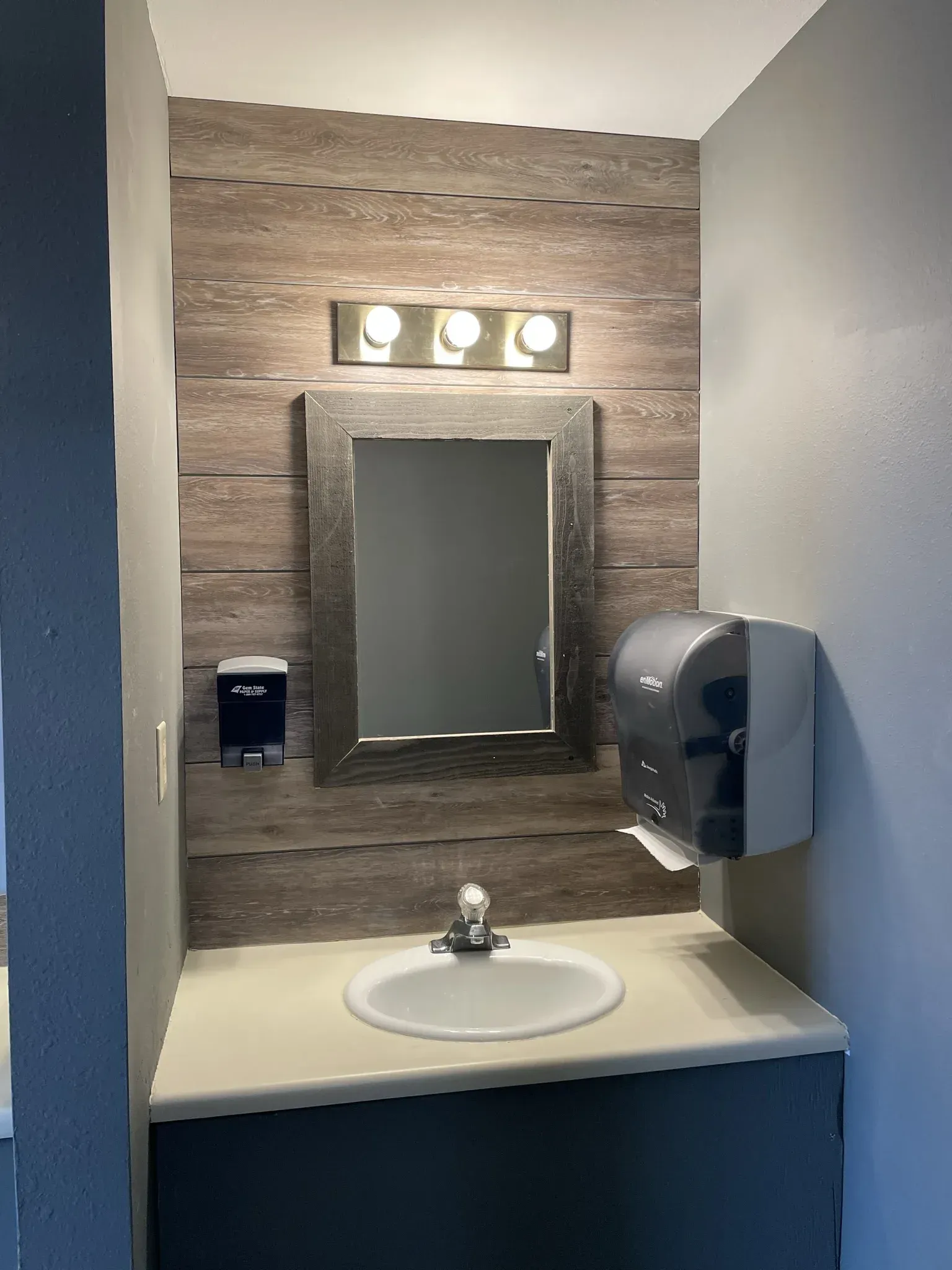 Bathroom with sink, mirror, soap dispenser, and paper towel dispenser. Wooden wall detail, gray walls.