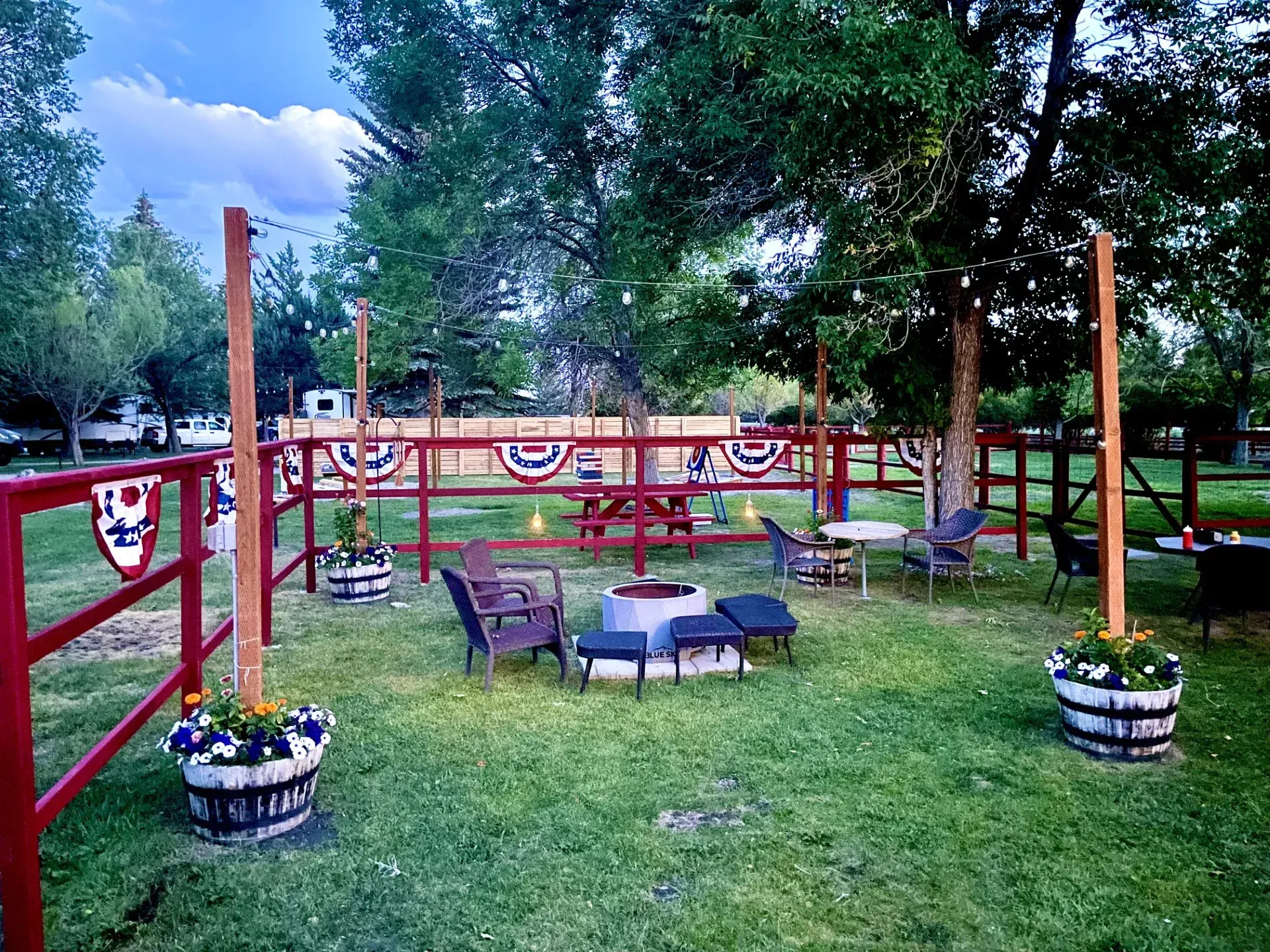Lawn with red fence, seating area, fire pit, and potted flowers. Trees and decorations suggest a celebration.