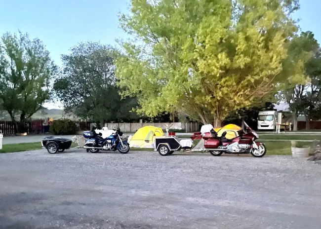 Motorcycles parked on gravel with yellow tents and trailer under trees, campsite setting.