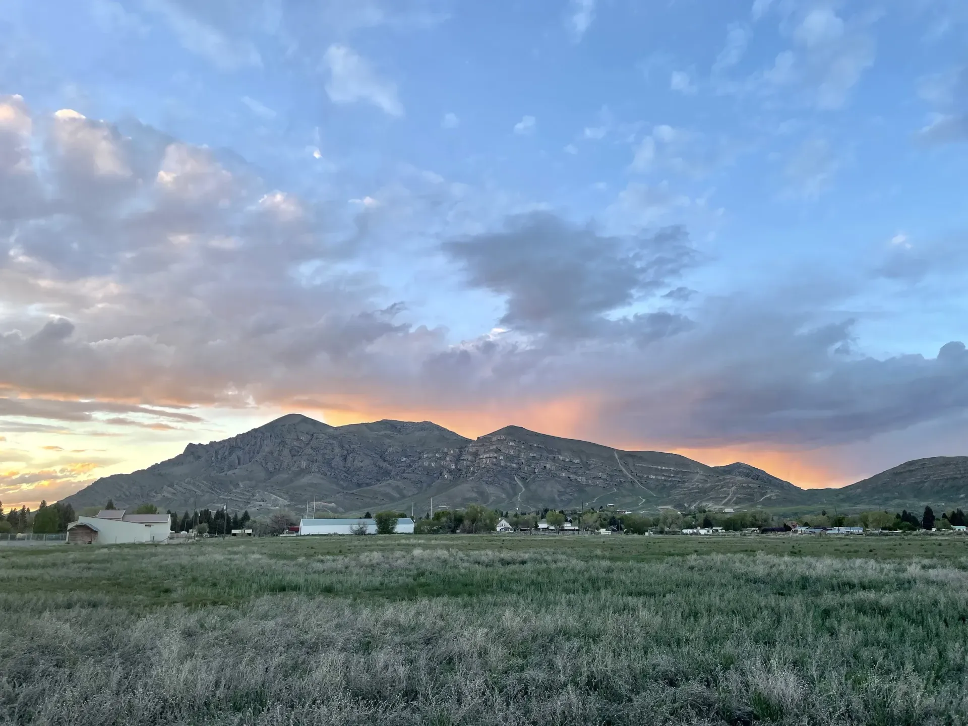 Mountain range under a cloudy, colorful sky at dusk, with buildings and a field in the foreground.