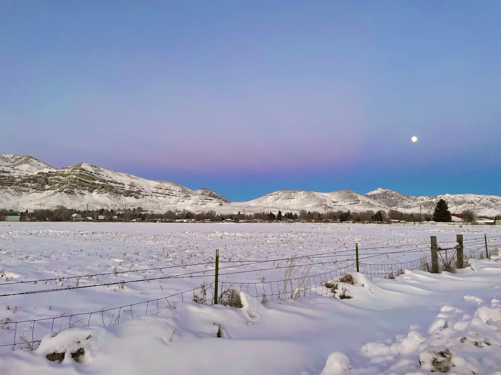 Snowy field with mountains under a colorful twilight sky. Moon visible.