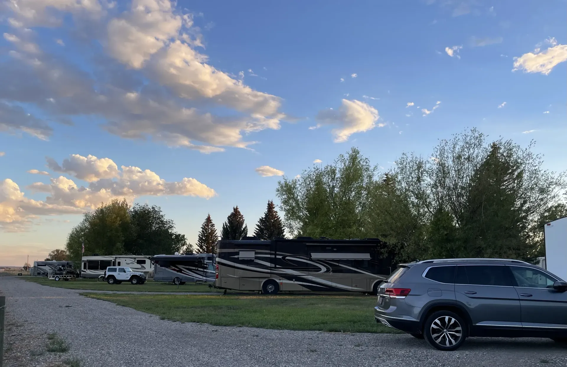 RV campground scene with motorhomes parked on gravel under a cloudy blue sky. A silver SUV is parked on the right.