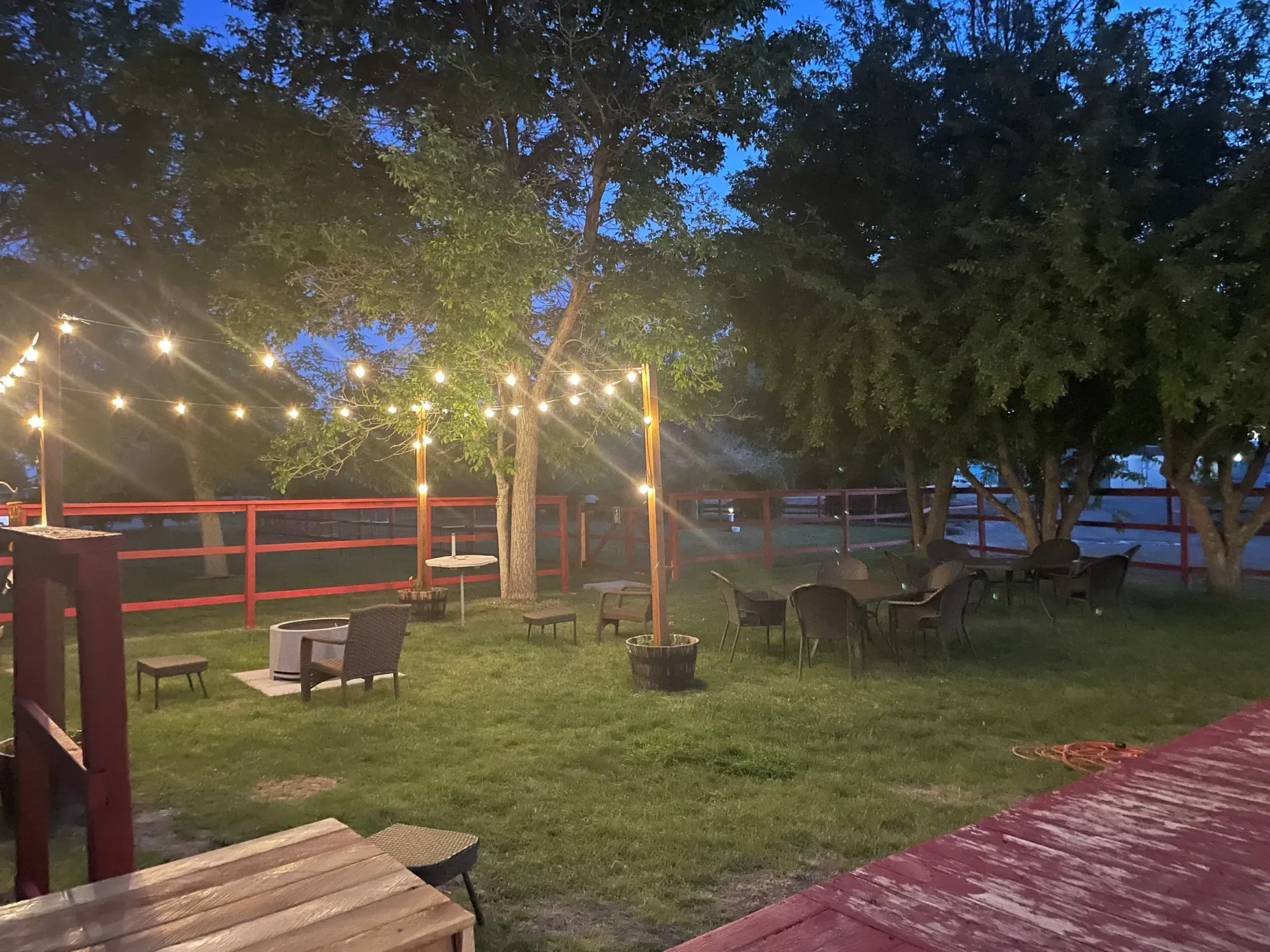 Nighttime outdoor seating area with string lights, trees, and fire pit on green grass, enclosed by a red fence.