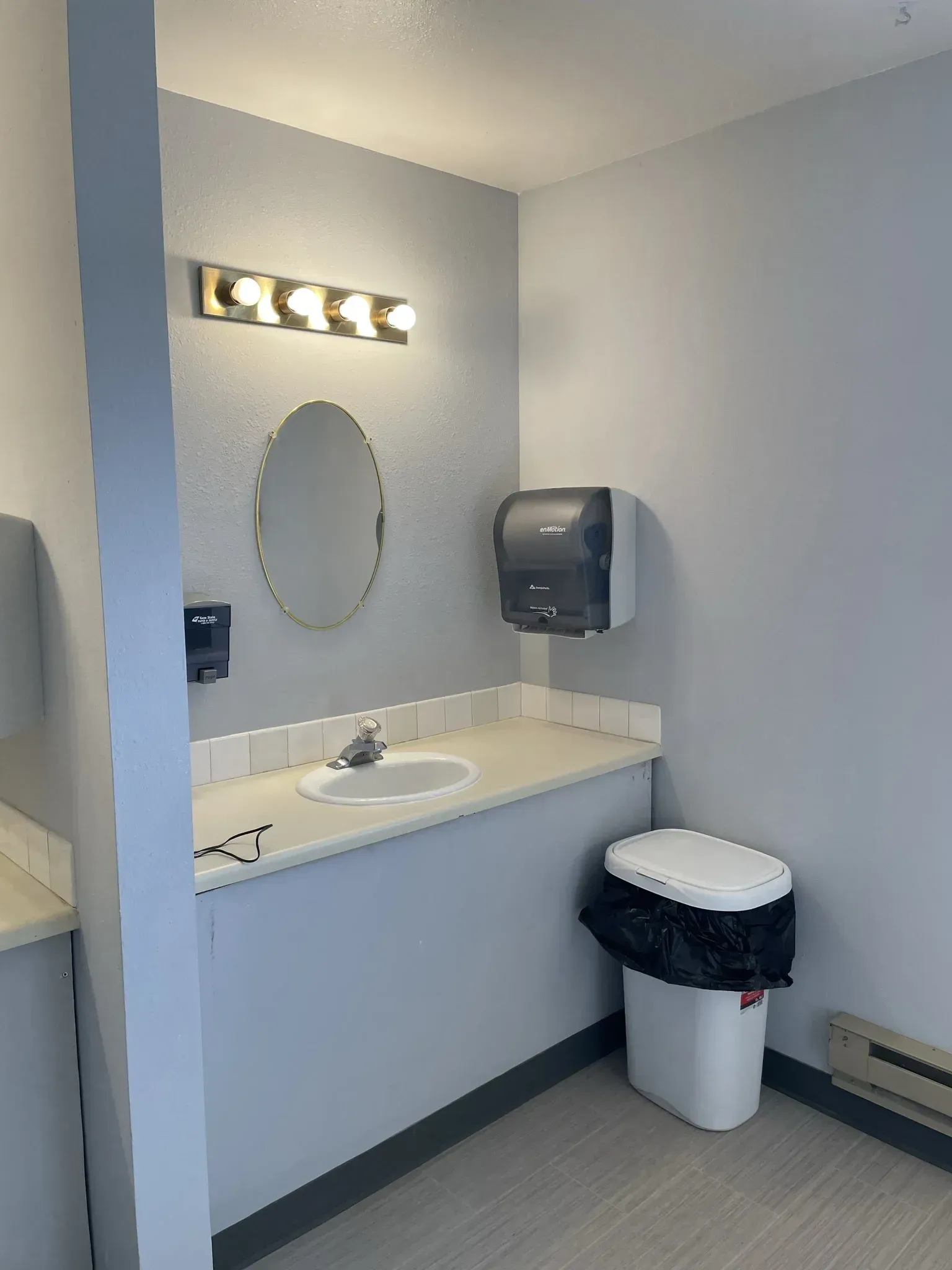Small, grey public restroom with a sink, mirror, and trash can.