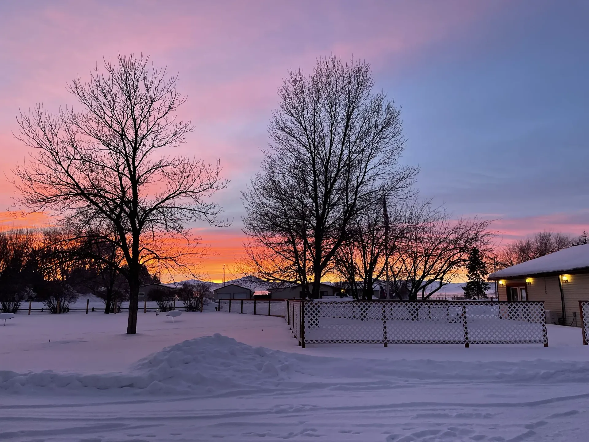Snowy winter scene with bare trees silhouetted against a vibrant orange and pink sunset sky.