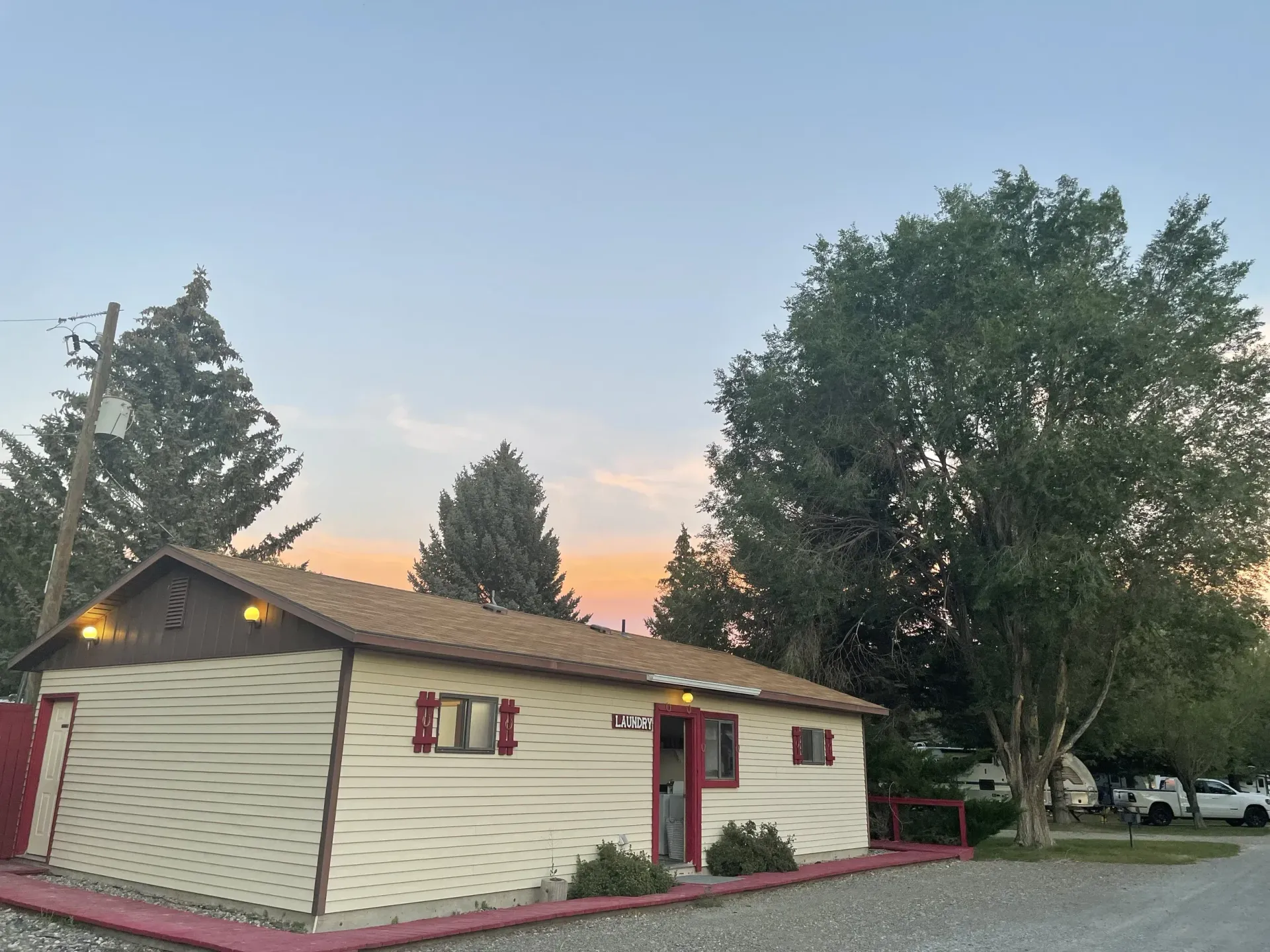 Small, light-colored building with brown roof and trim. Trees and a sunset sky in the background.
