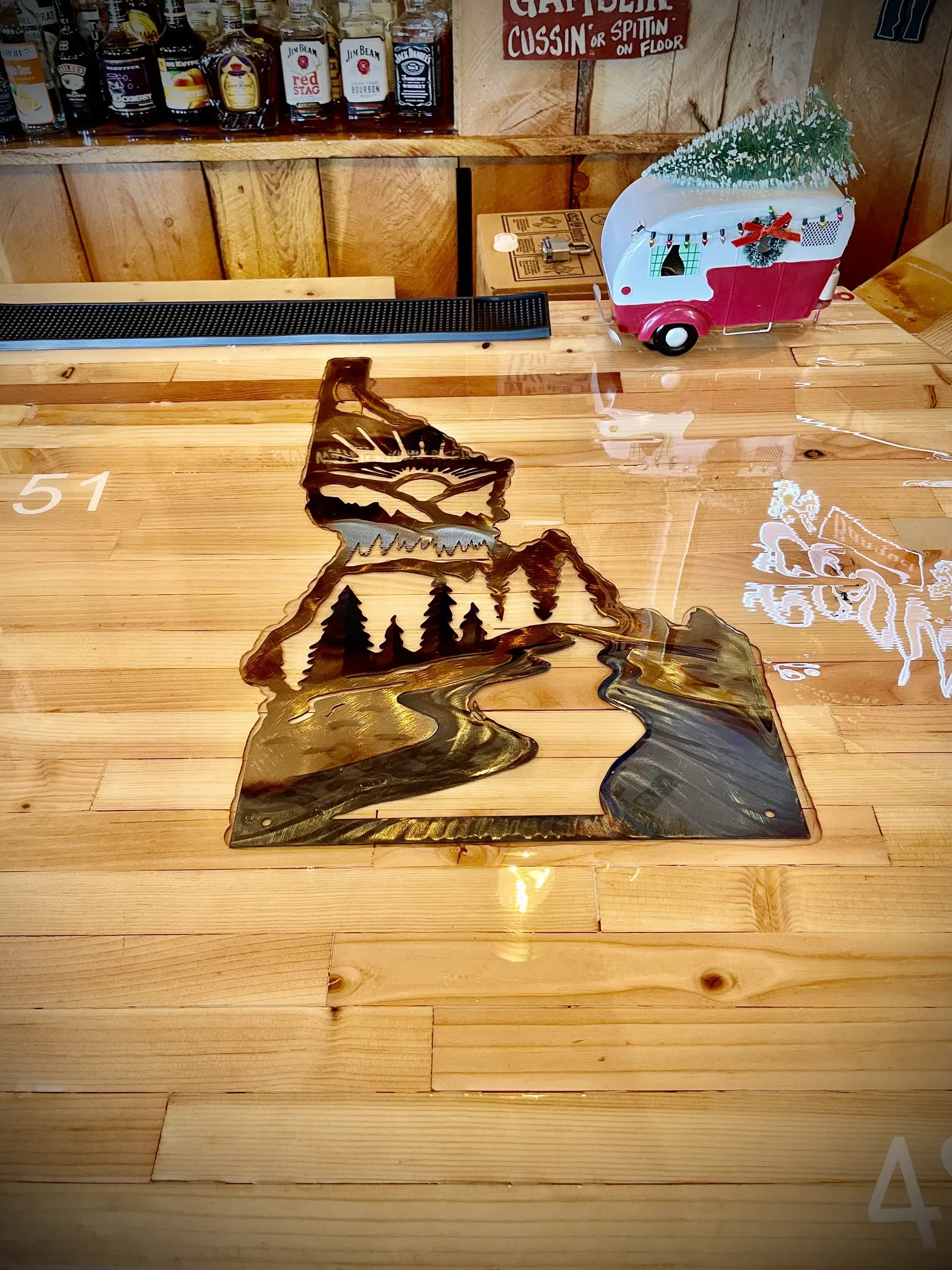 Metal mountain landscape art on a wood counter, with a miniature camper and bottles in the background.