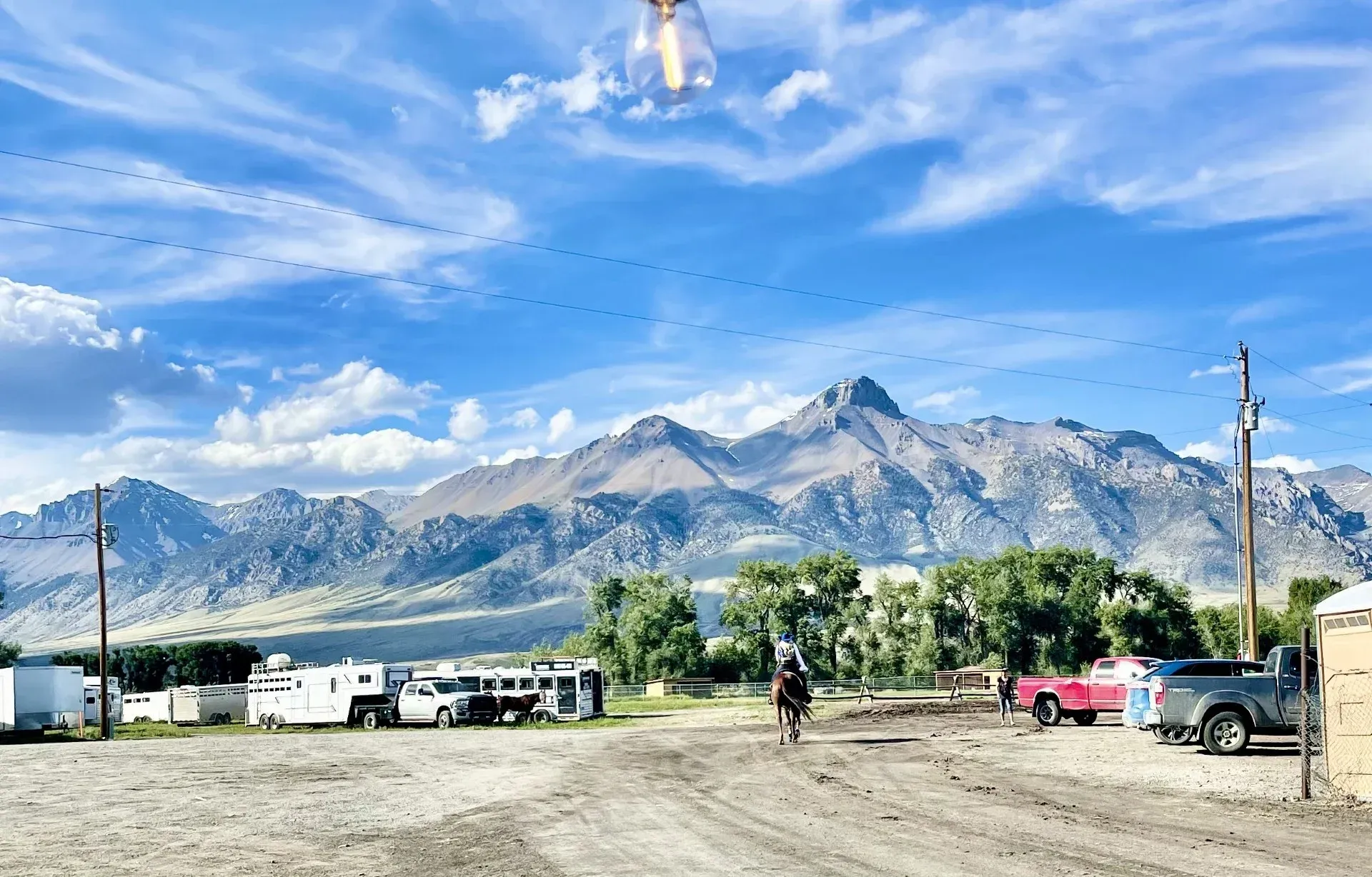A person on horseback rides towards mountains under a cloudy blue sky, with vehicles and buildings in the foreground.