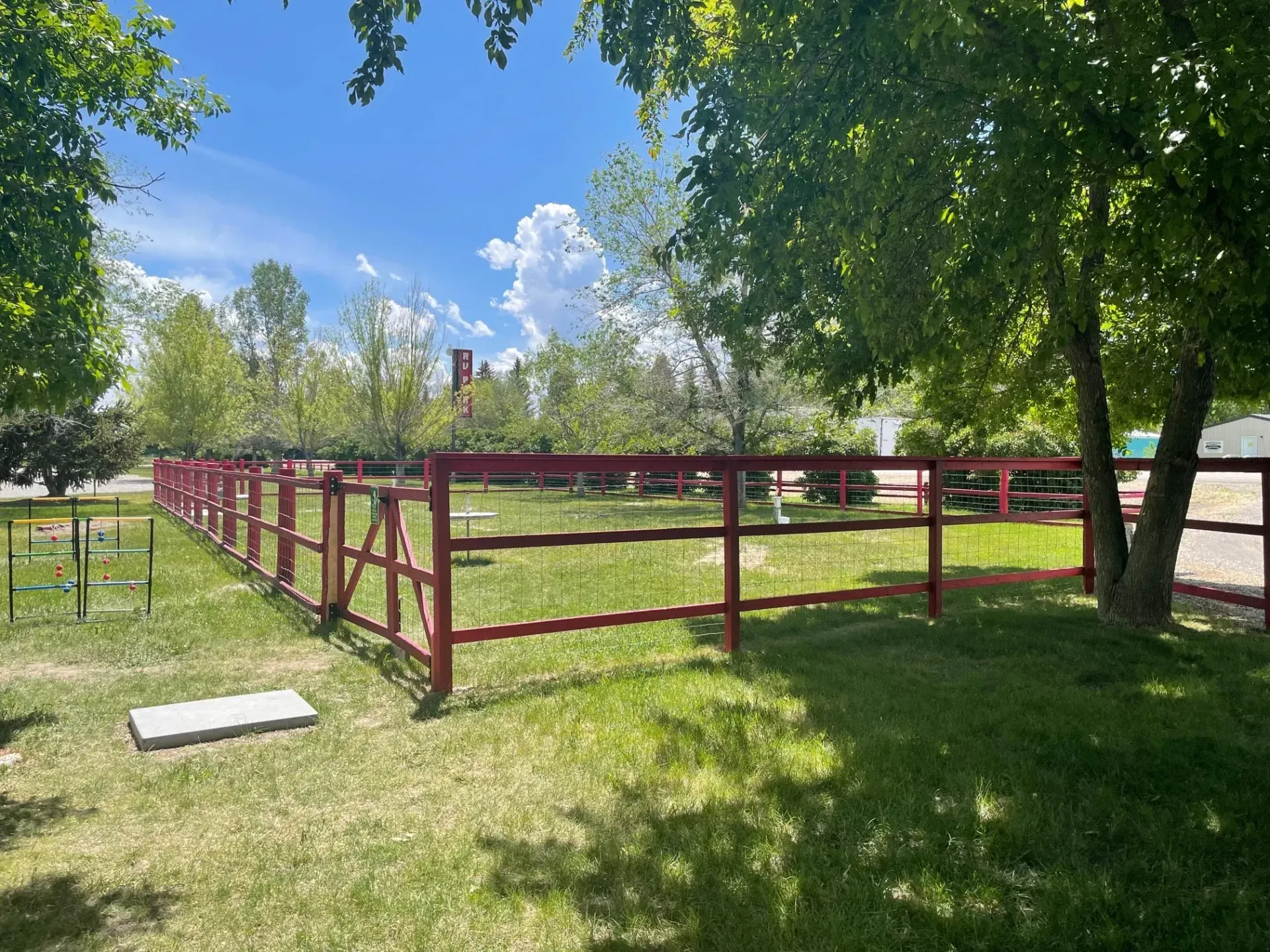 Red fence surrounding a grassy area, trees overhead, blue sky with clouds.