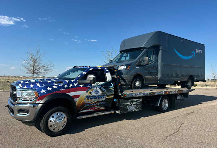 Tow truck with patriotic graphics carrying an Amazon delivery van on a sunny day.