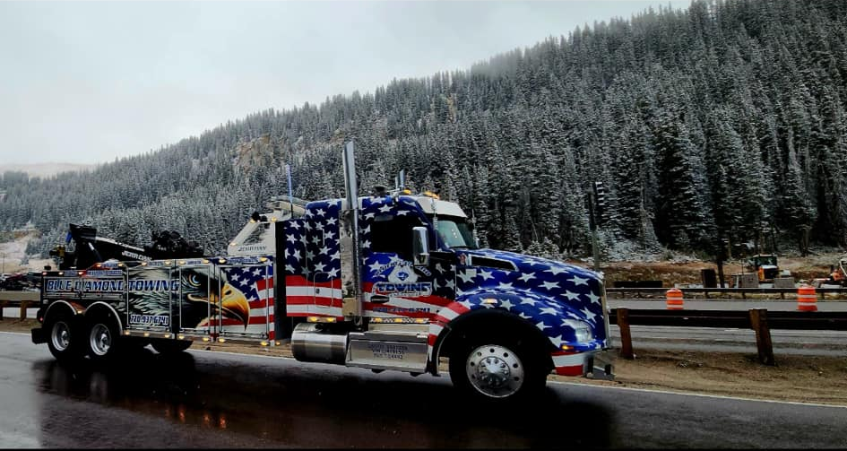 American flag-decorated tow truck parked on wet pavement near snow-covered mountains.