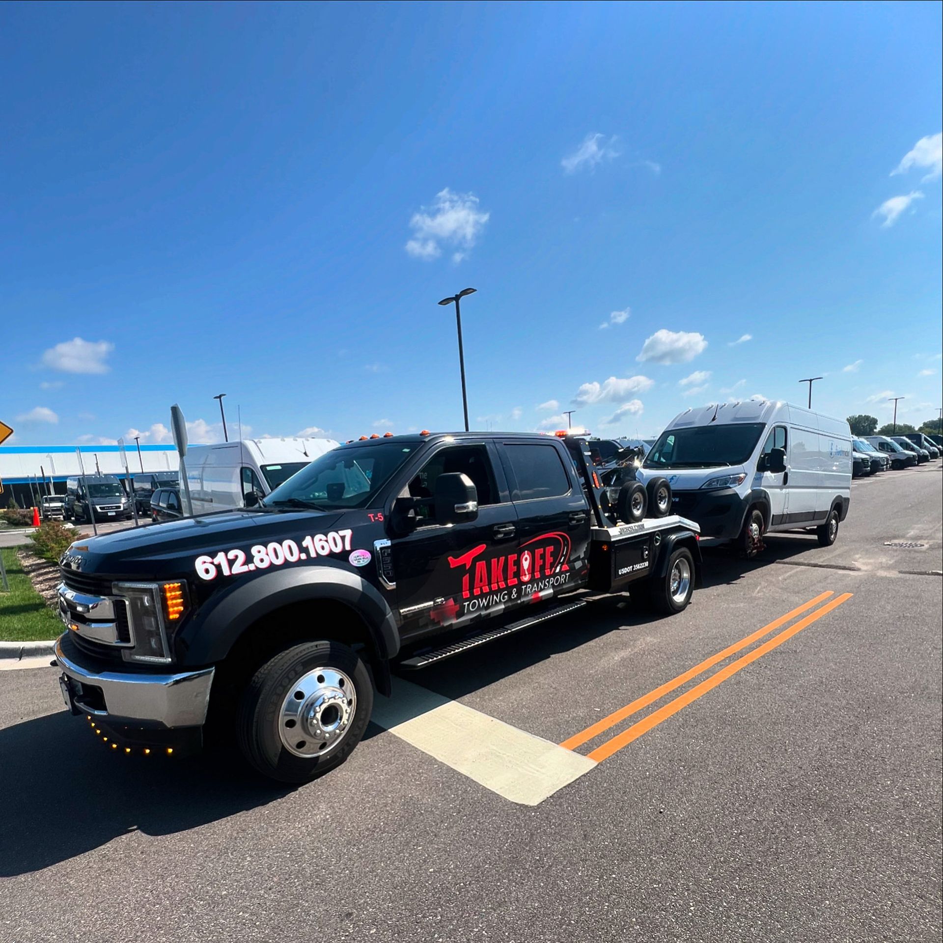 Black tow truck towing a white van on a road with a blue sky.
