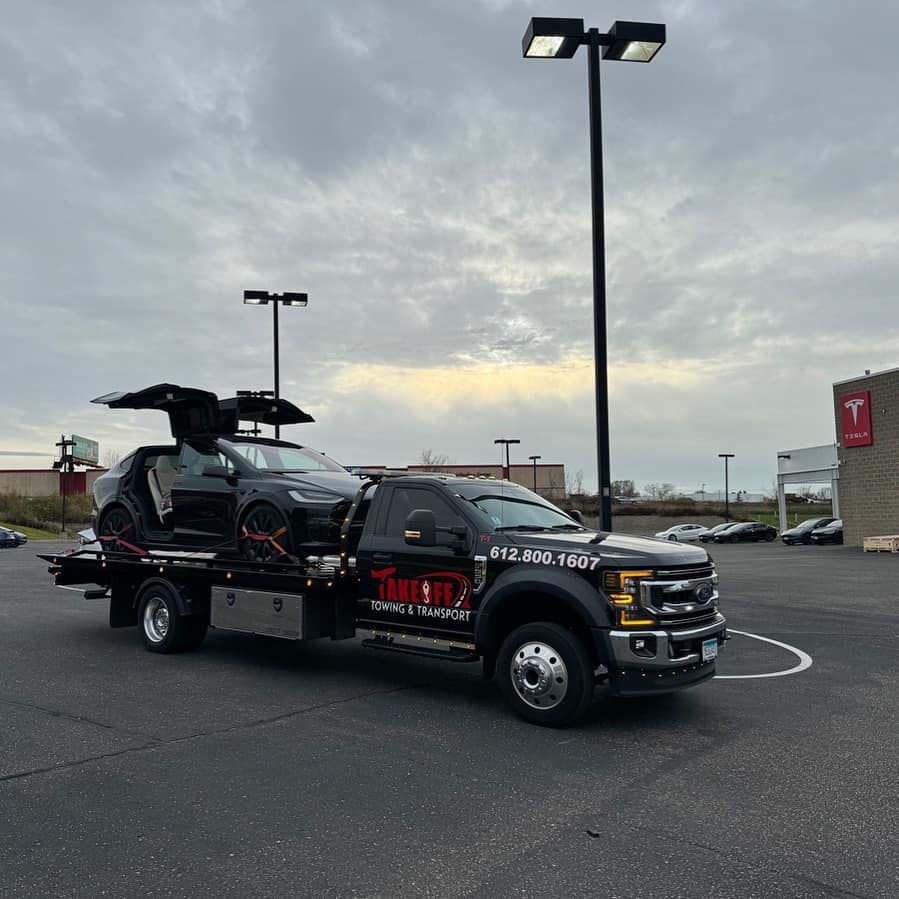Black Tesla SUV being towed on a flatbed tow truck in a car dealership parking lot. Cloudy sky overhead.