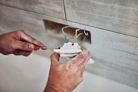 Electrician installing a wall outlet, using a drill beside exposed wiring and mounted cable boxes.