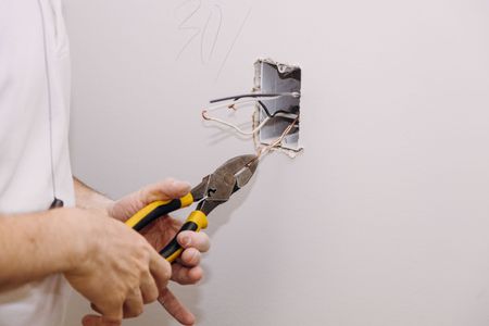 Hands using pliers to work on exposed electrical wires in a wall outlet opening