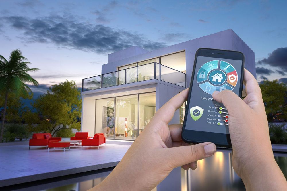 Mani che reggono uno smartphone mostrando i comandi della domotica davanti a una casa moderna al crepuscolo.
