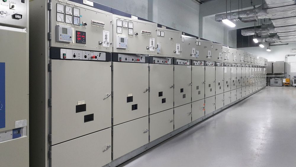 Lunga fila di pannelli di controllo elettrici grigi nel corridoio di uno stabilimento industriale.