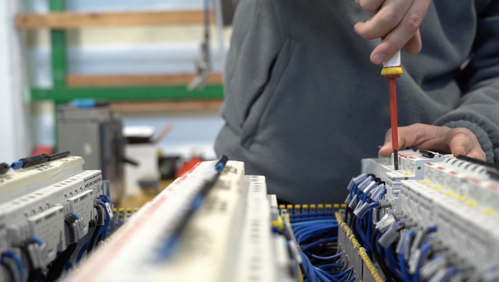 Una persona utilizza un cacciavite per lavorare su apparecchiature elettriche in un'officina, con fili e componenti ben visibili.