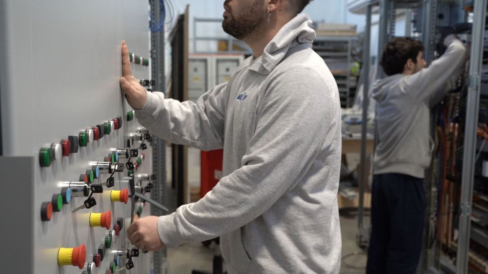 Tecnico al lavoro su un pannello di controllo in un impianto industriale, con pannelli e macchinari riflessi alle sue spalle.